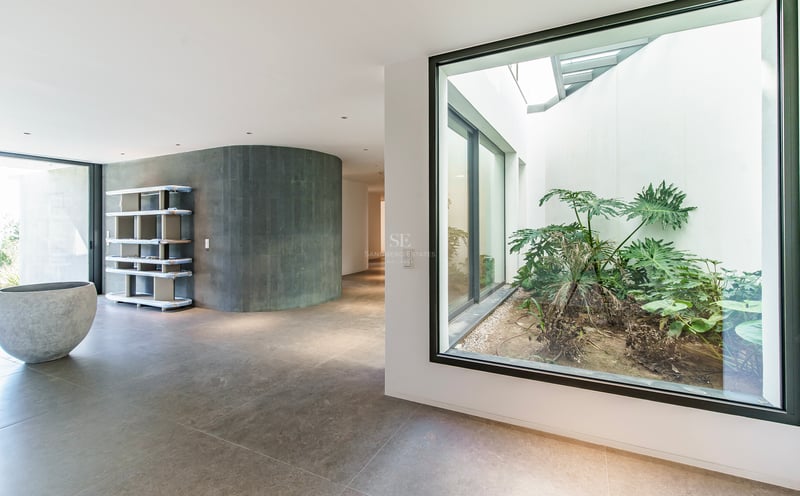 Couloir moderne avec un mur courbe en pierre grise et une grande fenêtre donnant sur un patio intérieur avec des plantes.
