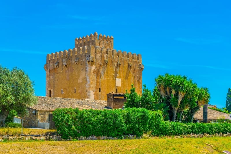 A grand medieval stone tower with battlements under a clear blue sky, surrounded by green hedges and trees.