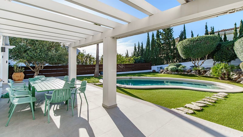 Skuggig terrass med grön matgrupp som vetter mot en pool och anlagd trädgård med cypresser.