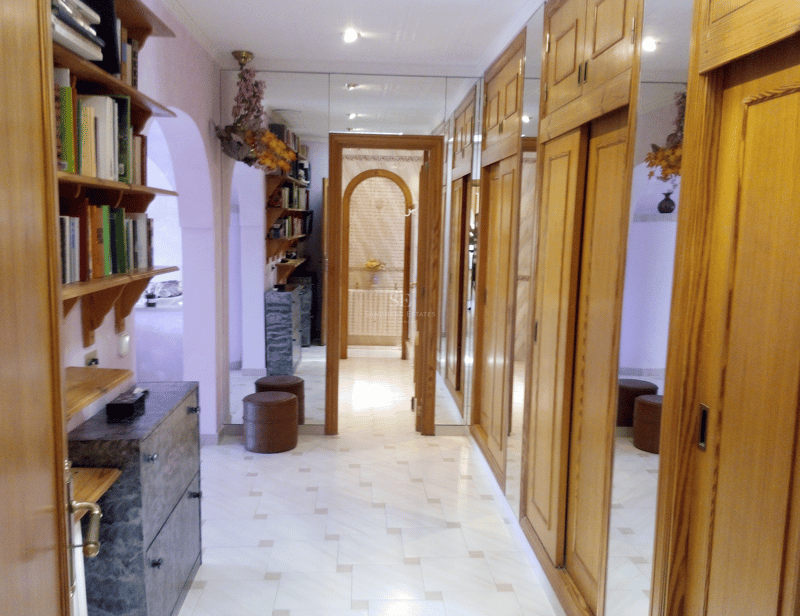 Pasillo con armarios de madera clara, puertas de espejo y suelo de mármol que conduce a un baño.