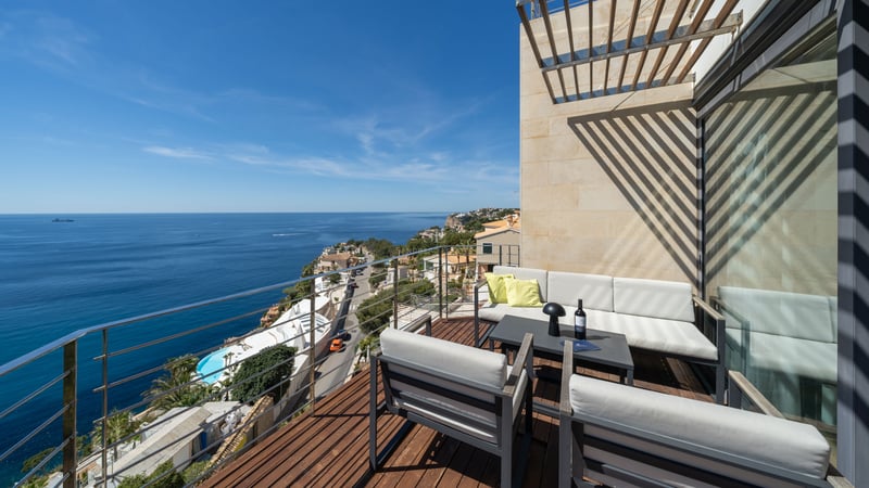 Wooden deck terrace with grey lounge furniture and glass railing overlooking the blue Mediterranean sea under a clear sky.