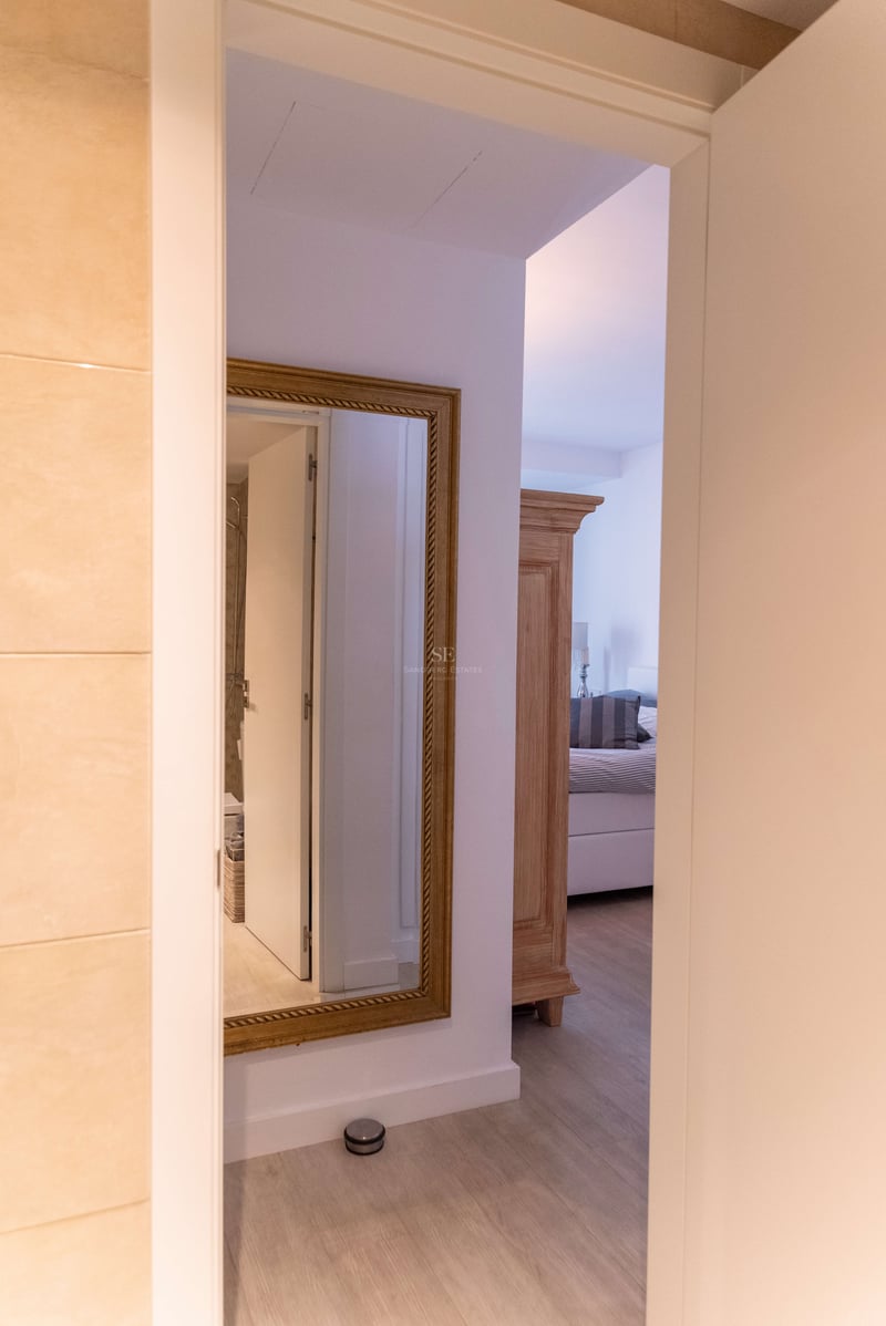 Blick durch eine offene Tür auf einen Flur mit goldgerahmtem Spiegel und Schlafzimmer mit Holzschrank.