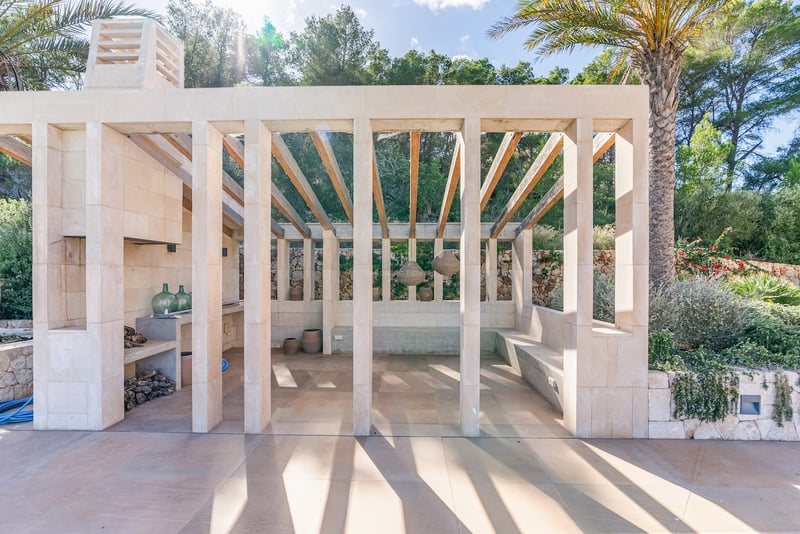 Pavillon moderne en pierre avec poutres en bois, cheminée extérieure et banquettes intégrées entouré de palmiers.