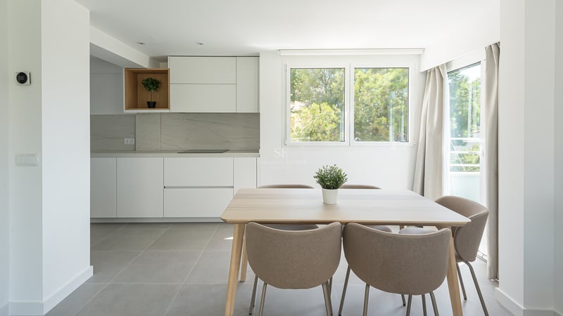 Salle à manger lumineuse avec table en bois clair, chaises taupe et cuisine blanche minimaliste vue sur jardin.