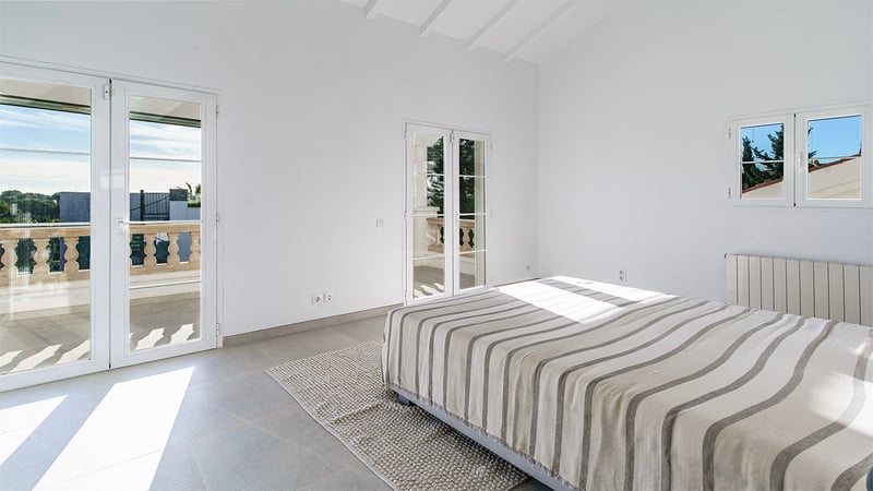 Minimalistiskt sovrum med randig säng, grått stengolv, högt tak med vita bjälkar och solig balkong.