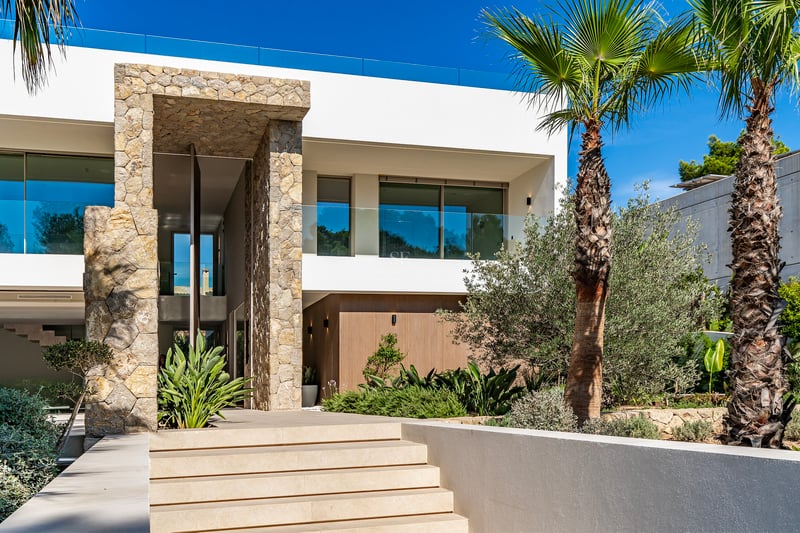 Entrée de villa moderne avec de larges marches en pierre, un haut portail en pierre et des palmiers.