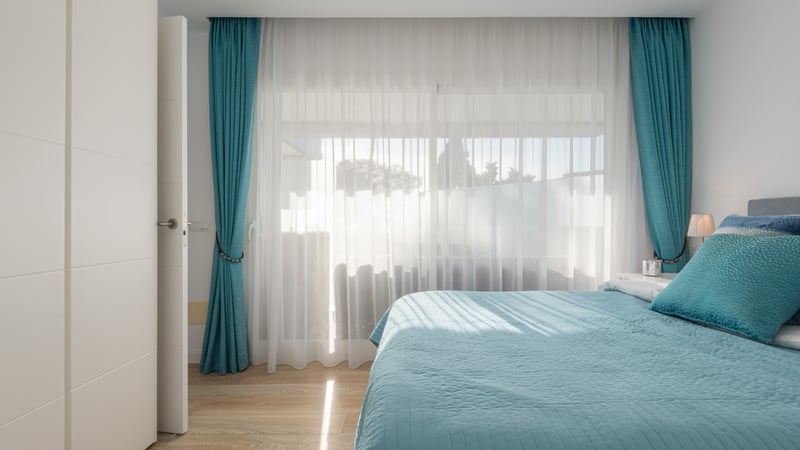 Chambre lumineuse avec lit turquoise, rideaux assortis et grandes fenêtres laissant entrer la lumière naturelle.