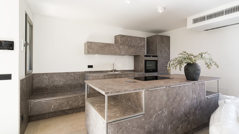 Modernt kök med stor köksö i gråmarmorerad sten, inbyggda vitvaror och minimalistiska vita väggar.