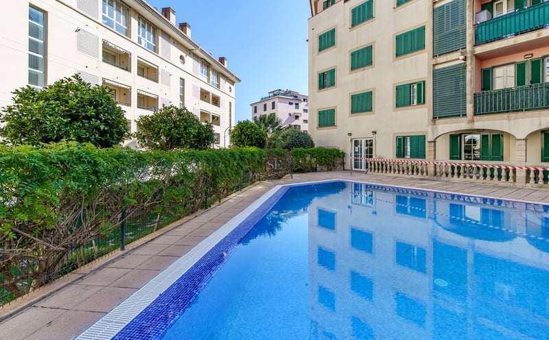 Utomhuspool med blå mosaik omgiven av stenplattor och byggnader med klassiska gröna fönsterluckor.