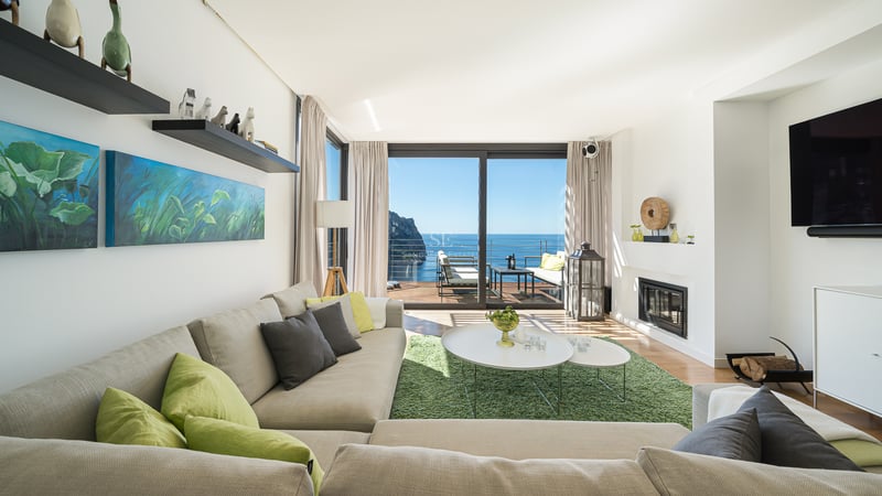 Bright modern living room with large glass doors opening to a sea-view terrace, featuring a neutral sectional sofa.