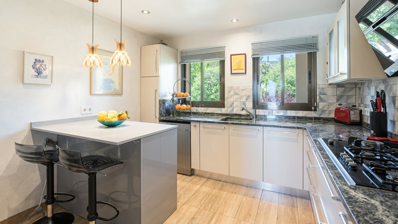 Modernt kök med vita skåp, bänkskivor i granit, köksö och fönster med utsikt över grönska.