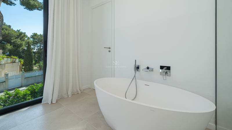 Baignoire îlot ovale blanche à côté d'une baie vitrée offrant une vue sur des pins méditerranéens luxuriants.