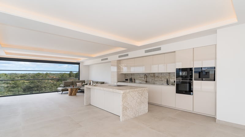 Minimalistische weiße Küche mit großer Marmorinsel, integrierten Geräten und indirekter Deckenbeleuchtung.