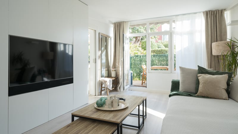 Modern living room with white wall unit, integrated TV, wooden coffee table, and glass door opening to a private garden.