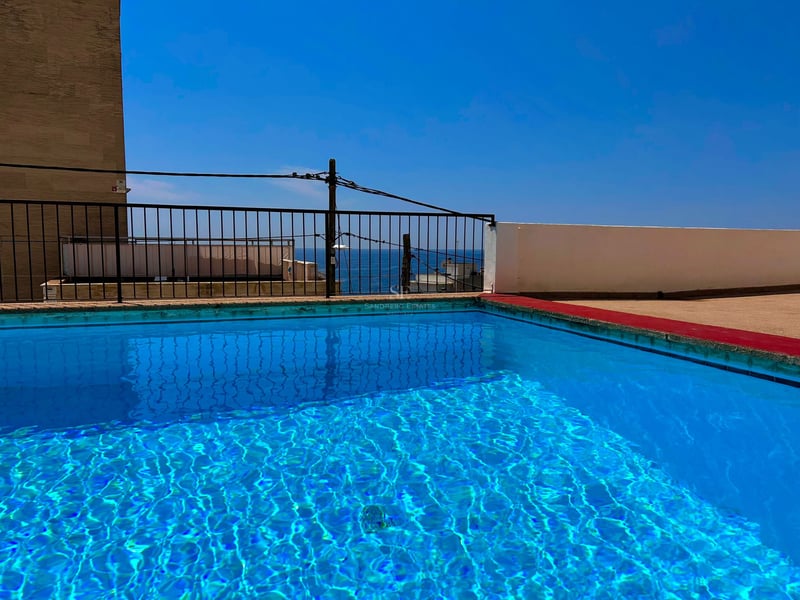Clear blue rooftop swimming pool with a black railing overlooking the sea under a bright blue sky.