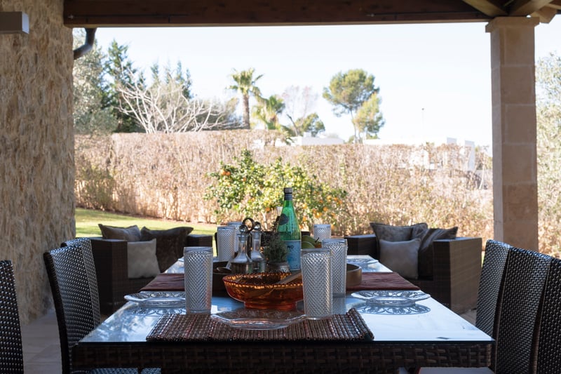 Övertäckt medelhavsterrass med matgrupp i konstrotting, naturstensväggar och utsikt över en lummig trädgård.
