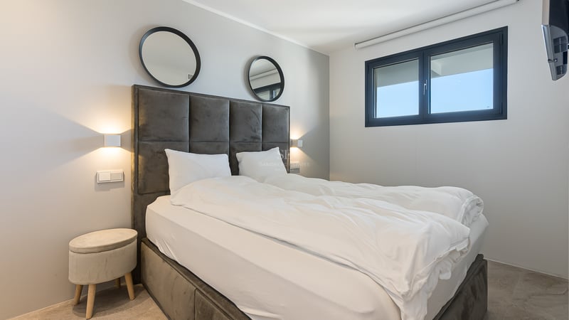 Modernes Schlafzimmer mit grauem Samtkopfteil, runden Spiegeln, Wandleuchten und schwarzem Fensterrahmen.