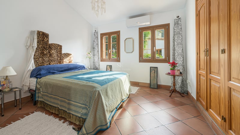 Sunny bedroom with terracotta tile floors, large wooden built-in wardrobes, and windows overlooking greenery.