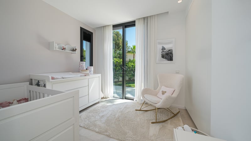 Chambre de bébé minimaliste avec berceau blanc, table à langer, fauteuil à bascule en bouclé et baie vitrée sur jardin.