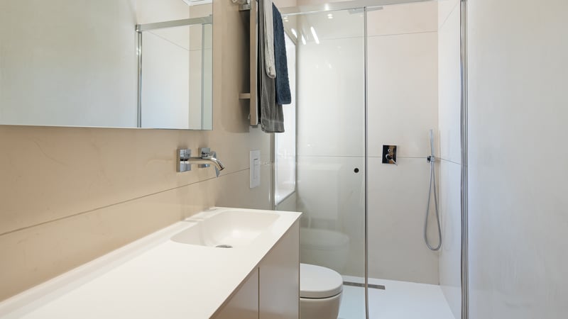 Modernt badrum med beige kakel, duschvägg i glas, vit kommod och kromade detaljer.