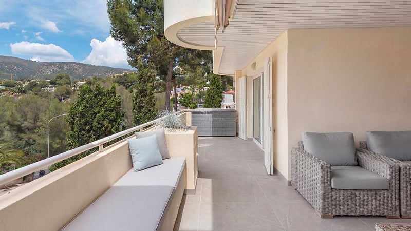 Modern covered terrace with grey rattan furniture, built-in bench seating, and views of lush green hills and pine trees.