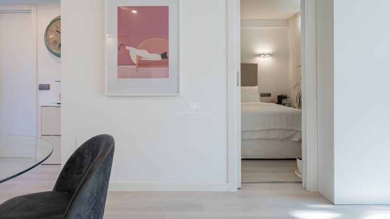 View through an open white door into a minimalist bedroom with a large bed, beige headboard, and warm wall lighting.