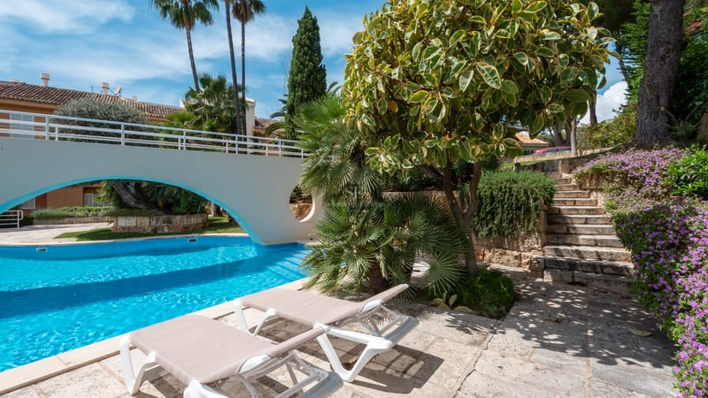 Sun loungers by a turquoise pool with a white arch bridge and lush landscaping.