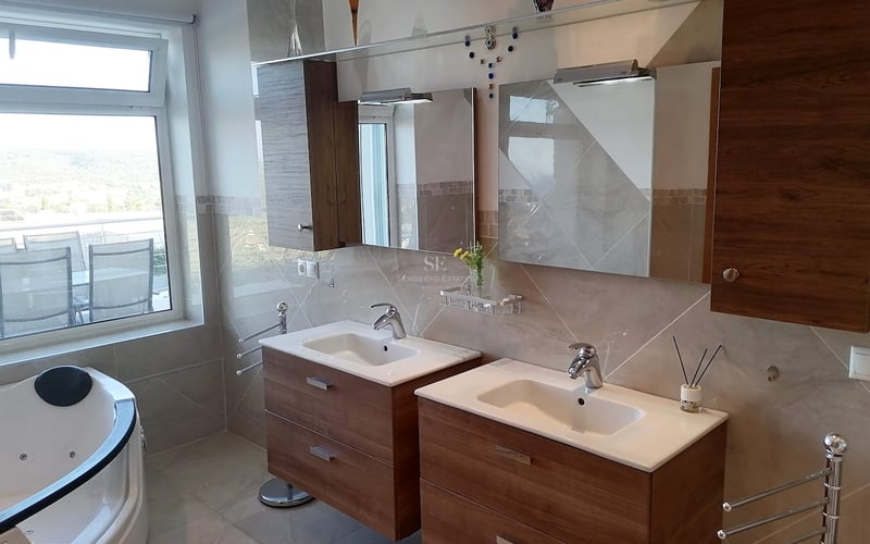 Double vasque avec meubles en bois, robinetterie chromée et baignoire balnéo près d'une grande fenêtre.