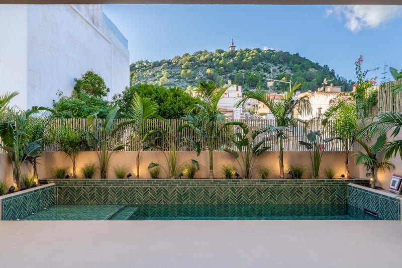 Piscine moderne avec carreaux verts en chevrons entourée de plantes tropicales et vue sur une colline sous un ciel bleu.