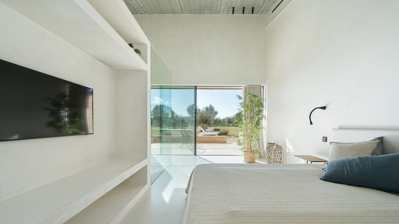 Minimalistiskt vitt sovrum med stora glaspartier som vetter mot en solig terrass och trädgård.