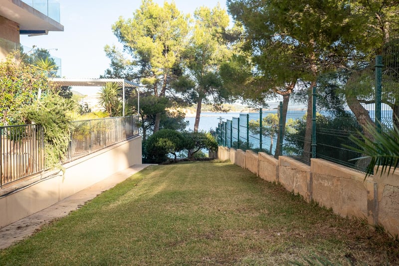 Gepflegter Rasen, umrahmt von Pinien und einem Metallzaun, mit Blick auf das türkisfarbene Mittelmeer.