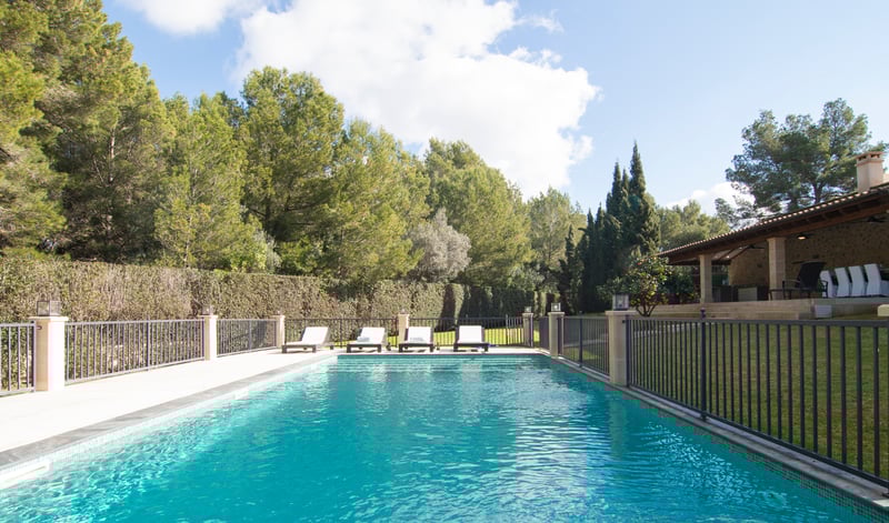 Rektangulär turkos pool med vita solstolar, omgiven av ett svart staket och lummiga tallar.