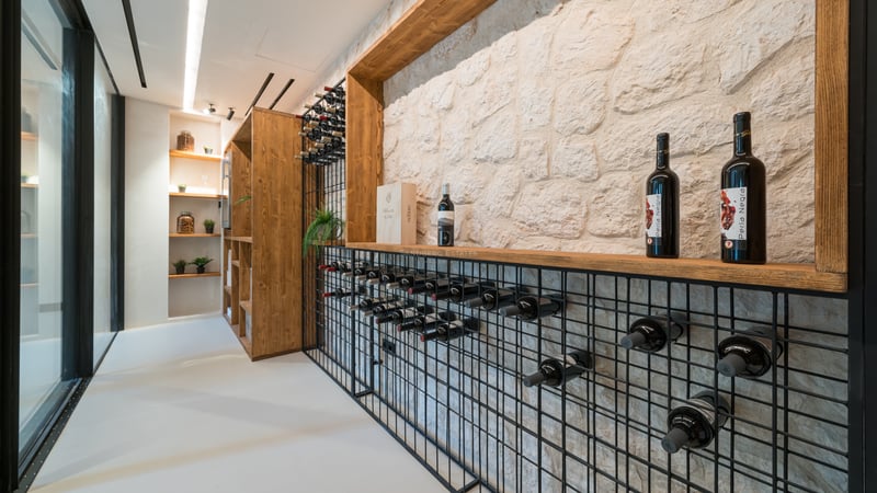 Modern vinkällare med vit stenmur, svarta metallställ för flaskor och detaljer i ljust trä.