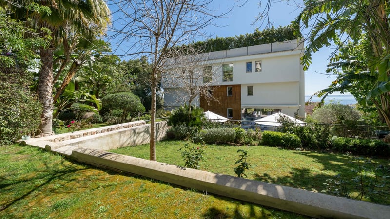 Moderne weiße Fassade mit Holzverkleidung, Blick auf einen grünen Garten und Poolbereich mit Sonnenschirmen.