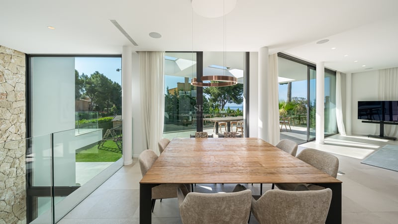 Grande table à manger en bois avec chaises beiges dans une pièce lumineuse avec baies vitrées et vue sur la mer.