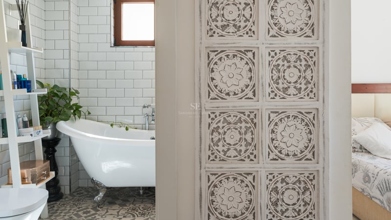 Salle de bain avec baignoire à pattes blanche, carrelage métro et sol à motifs, vue à travers une porte en bois sculpté.