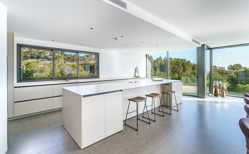 Cuisine lumineuse avec îlot blanc, tabourets en cuir et grandes baies vitrées donnant sur une forêt.