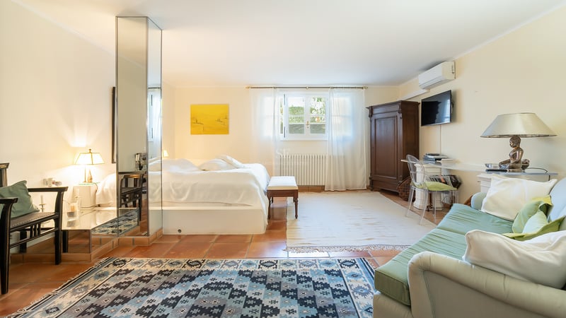 Grande chambre avec sol en terre cuite, pilier en miroir, literie blanche, armoire ancienne et coin salon.