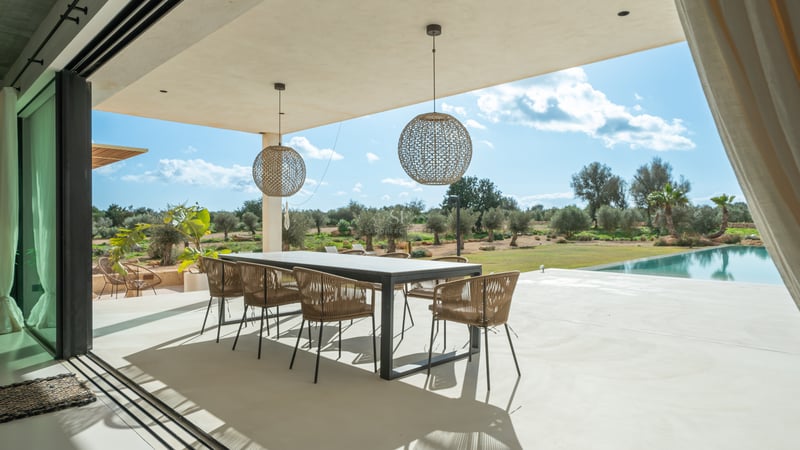 Övertäckt matplats utomhus med modernt bord och flätade stolar med utsikt över en trädgård med olivträd.