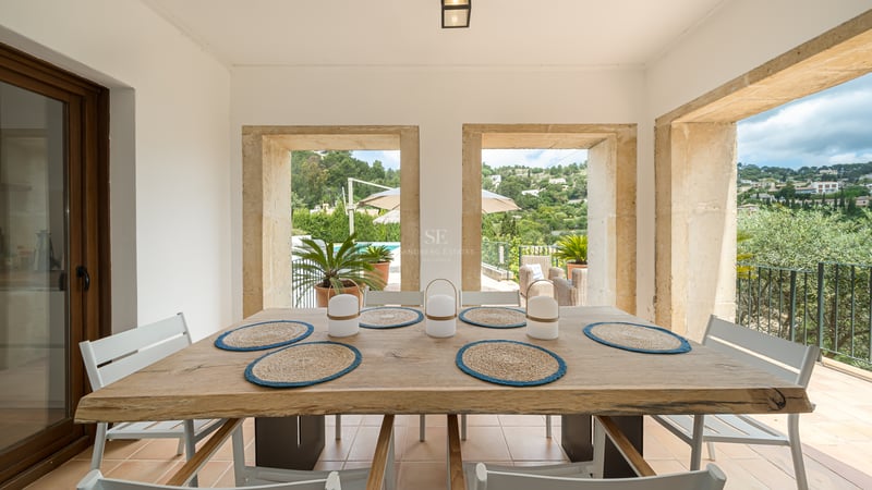Table à manger en bois face à des ouvertures en pierre offrant une vue sur un jardin verdoyant et une piscine.