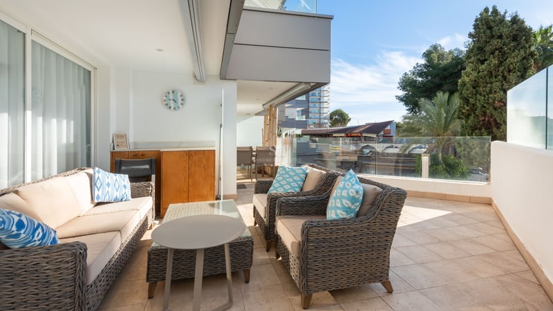 Grande terrasse avec canapés en osier gris, coussins bleus et garde-corps en verre sous un ciel bleu.