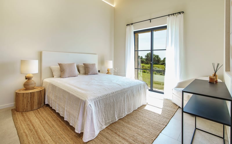 Helles Schlafzimmer mit weißem Bett, Jute-Teppich und bodentiefen Fenstern, die ins Grüne führen.