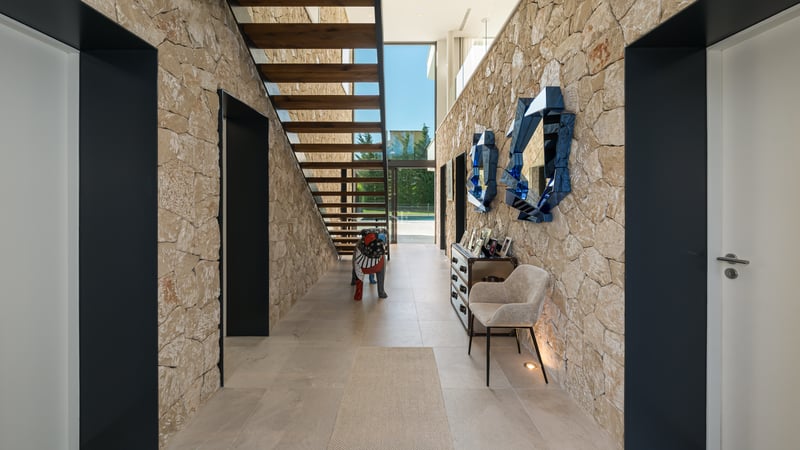 Couloir moderne avec murs en pierre, escalier flottant en bois et grandes baies vitrées vue sur le jardin.