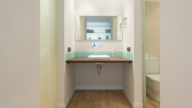 Badrum med svävande bänkskiva i trä, vitt fyrkantigt handfat, spegel och turkos glaspanel.