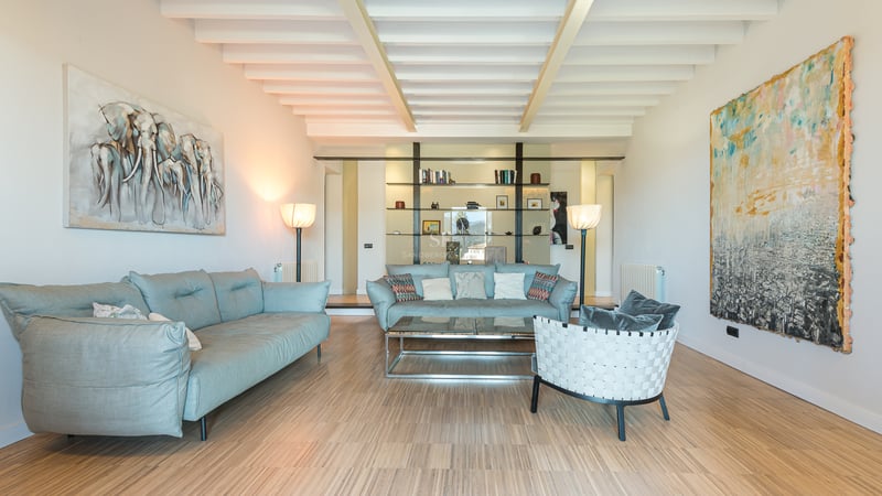 Rymligt vardagsrum med ljusblå soffor, vitt tak med bjälkar, trägolv och stora abstrakta målningar.
