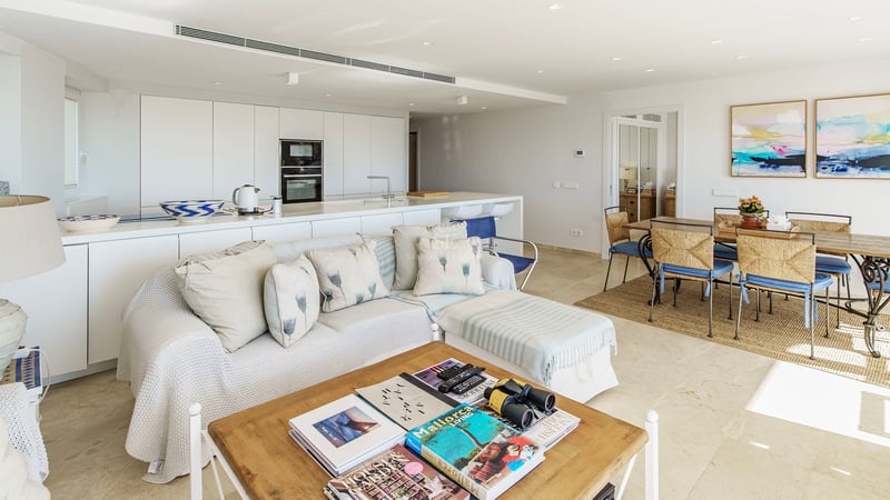 Bright open-plan living room with white sofa, wooden coffee table, dining set, and minimalist kitchen in the background.