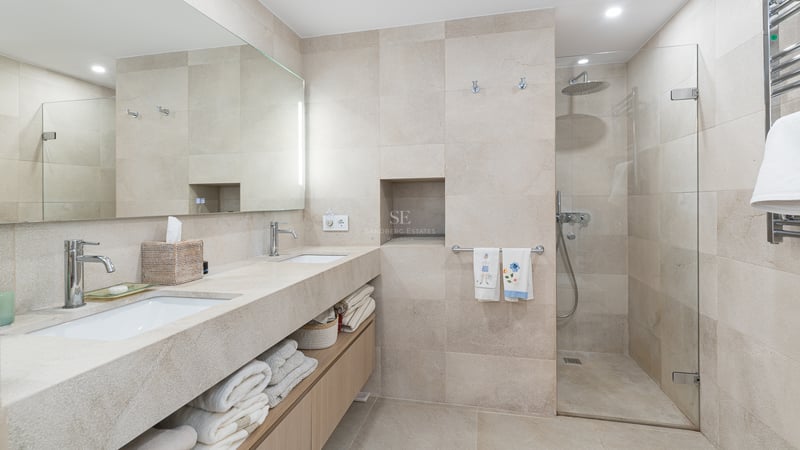 Modern bathroom featuring a double stone vanity, wood cabinets, and a walk-in glass shower with beige tiling.