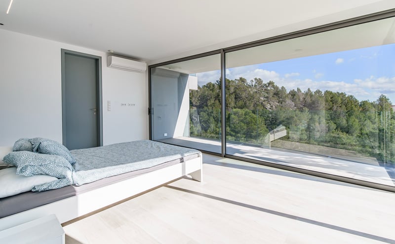 Chambre lumineuse avec baies vitrées ouvrant sur un balcon surplombant une forêt verdoyante.