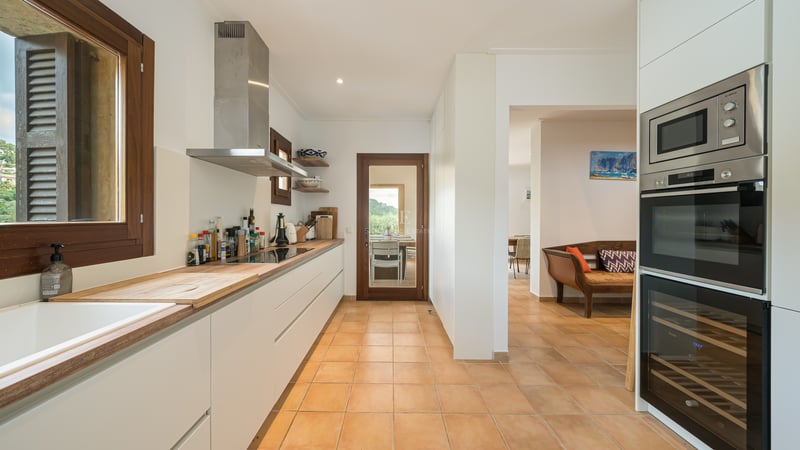 Cuisine blanche moderne avec plans de travail en bois, sol en terre cuite et hotte aspirante en acier inoxydable.