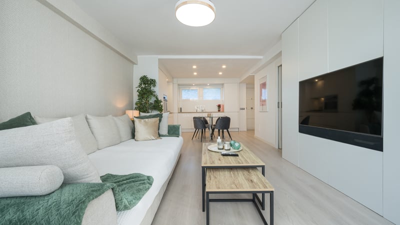 Bright modern living room featuring a white sofa, light wood floors, and a large flat-screen TV integrated into white cabinetry.
